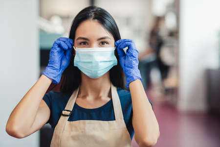 Epidemic prevention. Young female haordresser putting on protective mask before clients visit at...