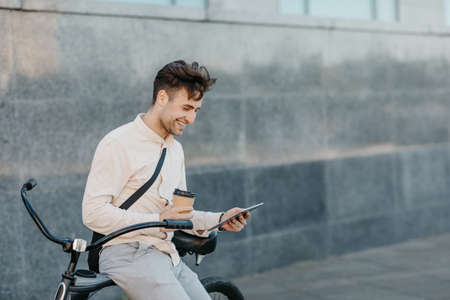 Coffee take away and online outdoors. Happy man in business clothes holds cup, looks at tablet and sits on bicycle on street, free spaceの写真素材