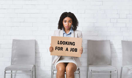 Serious African American woman holding sign with words LOOKING FOR JOB at company lobby, panoramaの写真素材