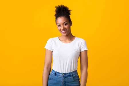 Portrait of cute african american girl looking at camera with shy charming smile over yellow wall