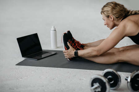 Sporty woman in sportswear sitting on floor with dumbbells and bottle of water or protein shake and, using laptop for workout at home in living roomの写真素材