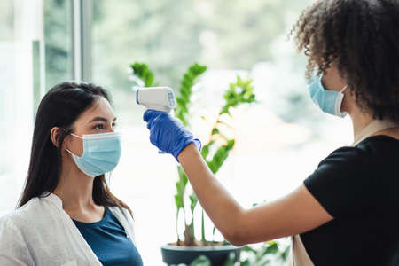 Woman checking temperature to client using infrared tool in beauty salon. Social distancing concept