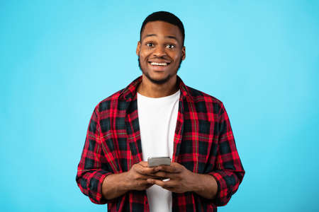 Cheerful Black Guy Using Smartphone Smiling To Camera Standing Over Blue Background. Studio Shot