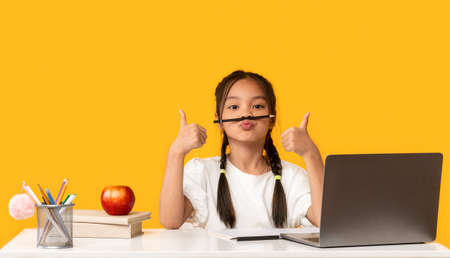 Funny Asian School Girl At Laptop Gesturing Thumbs-Up Having Fun Posing Sitting At Desk In Studio On...