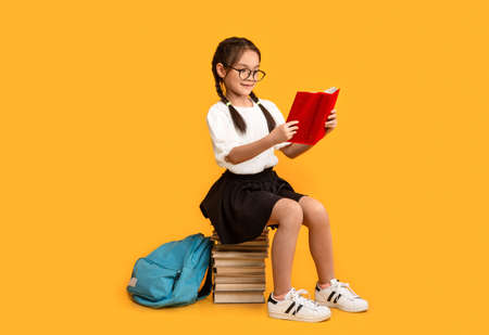 Cheerful Asian Schoolgirl Reading Sitting On Book Stack Learning Over Yellow Background In Studio. School Education Conceptの写真素材