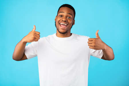 Black Millennial Guy Gesturing Thumbs Up With Both Hands Smiling To Camera Standing Over Blue...