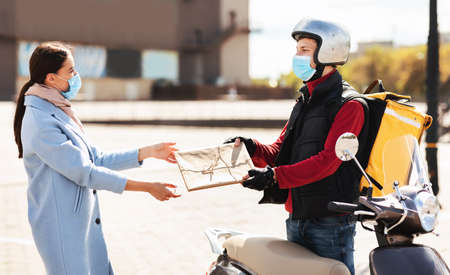 Safe Delivery Service. Courier In Protective Mask Delivering Package To Customer Standing With Scooter In Urban Area Outdoor.の写真素材