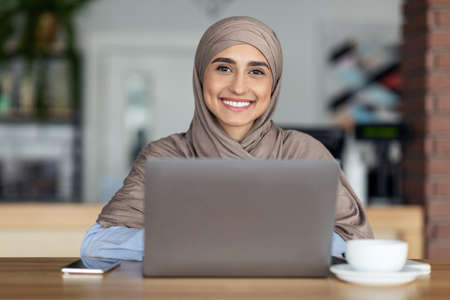 Cheerful muslim woman in hijab working online at cafe, using laptop and drinking coffeeの写真素材