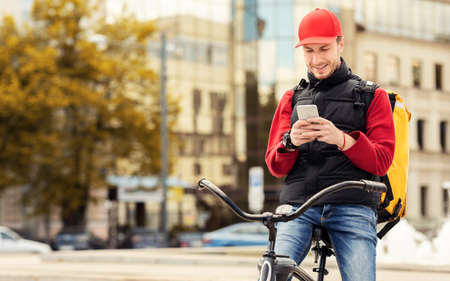 Courier Using Delivery Application On Smartphone Delivering Restaurant Food On Bicycle In City. Free Space For Textの写真素材