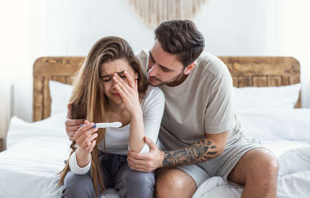 Calm down in difficult times and problems with conception. Sad man hugs crying woman and looks at pregnancy test in bedroom interiorの写真素材