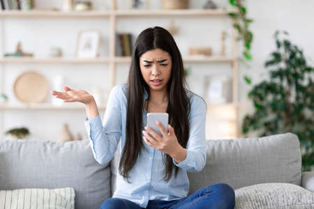 Frustrated Asian Girl Sitting With Smartphone On Couch, Emotionally Reacting To Message, Received...
