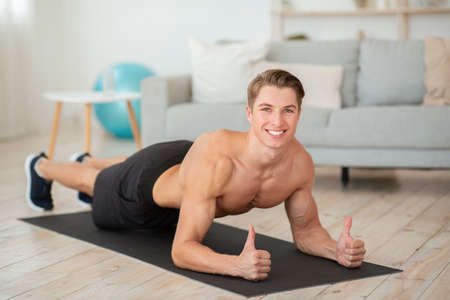 Strong man in cozy atmosphere working on muscles and fit body. Smiling guy with muscular torso makes plank and shows thumb up in living room interiorの写真素材