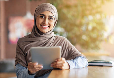 Happy girl in hijab with digital tablet smiling at camera, sitting at cafe alone, copy space