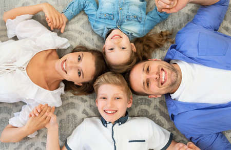 Family Bond. Happy Parents And Kids Lying Holding Hands Smiling To Camera Posing On Floor Indoors....