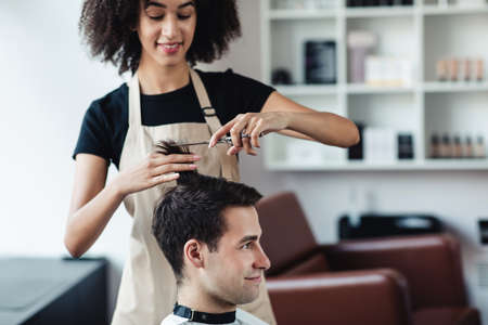 New hairstyle. African american hairdresser cutting hair for young man at barber shop, free spaceの写真素材