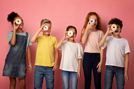 Diverse schoolkids holding yummy donuts in front of their faces on pink backgroundの写真素材