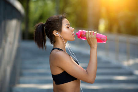Closeup of african fitness model drinking water during training at park, side view