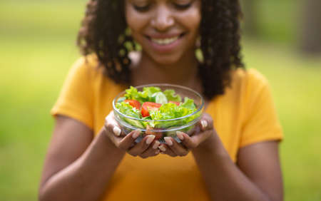 Healthy eating concept. Happy black girl holding bowl of fresh vegetable salad outdoors, selective...