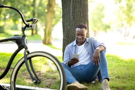 Millennial black man with bike sitting near tree and using mobile phone at green summer parkの写真素材