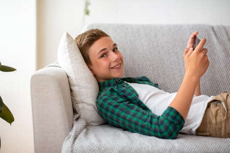 Side view of positive teen guy gaming on smartphone while resting at homeの写真素材