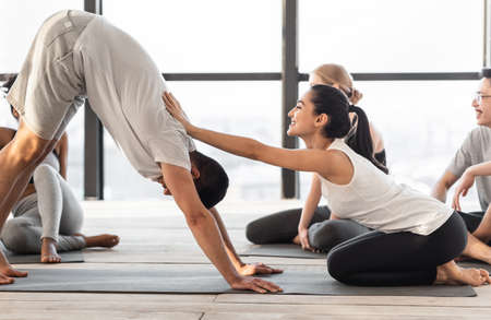 Female coach correcting yoga beginner at group class training in studio, side view with copy space