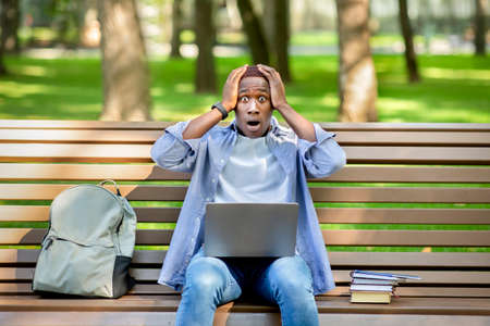 Big failure. Young black guy holding his head in panic near laptop computer at city park
