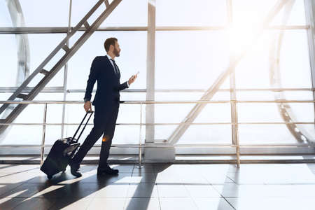 Businessman with smartphone and luggage walking by airport, looking through the window, sun glare, copy spaceの写真素材
