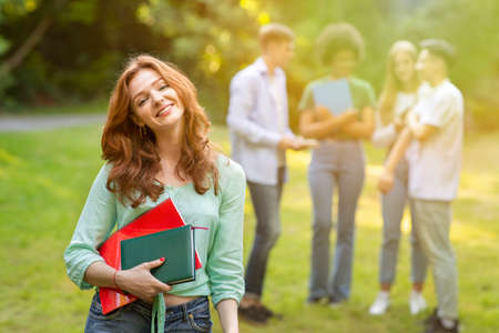 Cute young student girl with workbooks posing outdoors with her classmates on background, resting at...