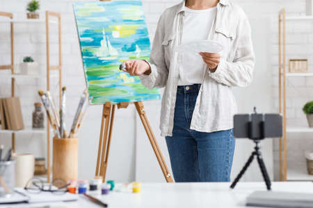Painting and modern technology. African american girl with brushes and paints painting at home and...