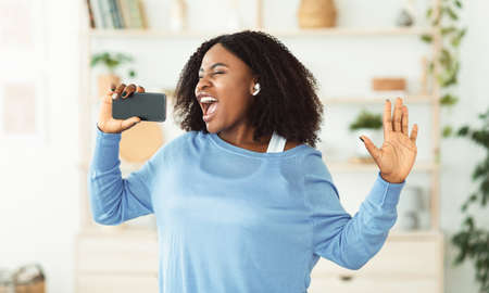 Carefree Singer. Portrait of african lady singing and using cell phone as mic, dancing and wearing...