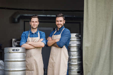 Own small business. Two handsome guys in aprons crossed arms over chest and stand near metal barrels in brewery warehouse, free spaceの写真素材