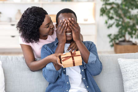 Black woman giving her boyfriend birthday gift at home, man covering his eyes with palms, copy spaceの写真素材
