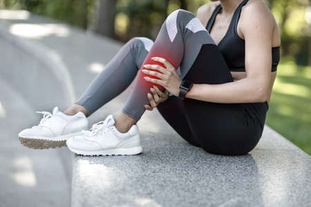 Unrecognizable black woman in sportswear rubbing her leg, sitting on stairs at park, sport injuries conceptの写真素材