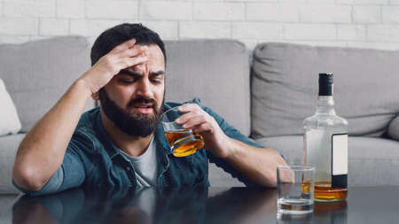 Problem of alcohol dependence. Sad man with beard looks at glass of whiskey, near on table are...