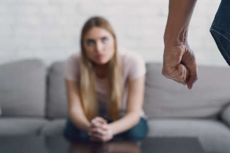 Fear and problems in family. Angry man with fist close up, and sad woman sits at sofa in living room interiorの写真素材