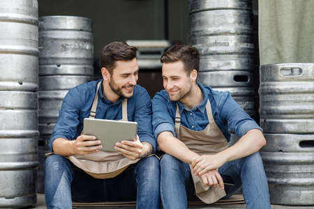 Friends and startup. Two handsome guys in aprons talking and looking at tablet, near metal barrels...