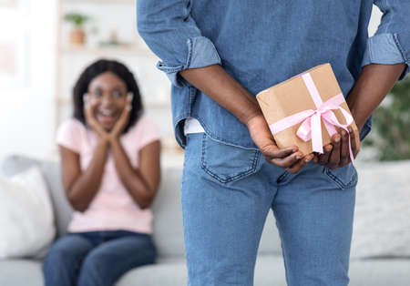 Man holding present behind back for his surprised girlfriend, celebrating anniversary at homeの写真素材