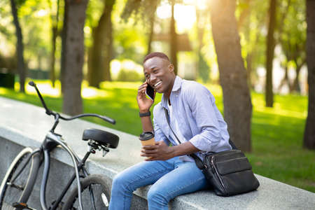Confident African American man with bicycle and coffee having conversation on mobile phone at city...