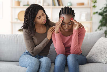 Black girl comforting her sad friend, sitting together on couch at home, friendship conceptの写真素材