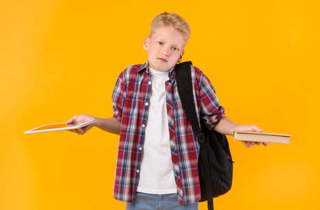 Gadgets Or Books. Unsure Blonde Schoolboy Comparing Tablet And Book Over Yellow Wall, Looking At...