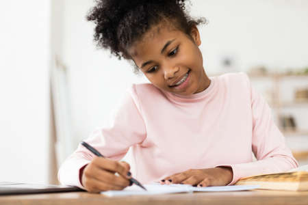 African Teen Girl Doing Homework Writing In Exercise-Book Sitting At Desk At Home. Selective Focus