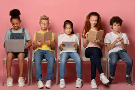 E-learning concept. Focused diverse schoolkids with gadgets and study materials sitting on chairs against pink backgroundの写真素材
