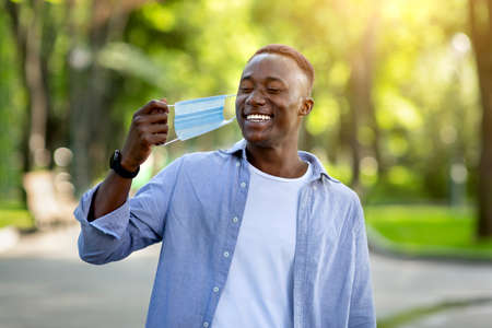 Coronavirus epidemic is over. Smiling black man removing his medical mask at city park