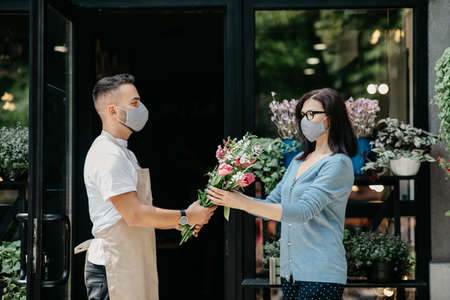 Customer service in flower shop. Man in apron and protective mask gives bouquet to client during quarantine, indoors