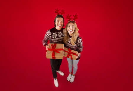 Happy couple wearing woolen sweaters and deer horns with Christmas gift over red backgroundの写真素材