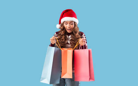 Big Christmas sale. Surprised young lady in Santa hat peeking into gift bags on blue background