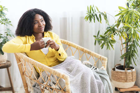Peaceful Morning. Calm black girl drinking hot coffee, sitting in wicker chair with blanket and...