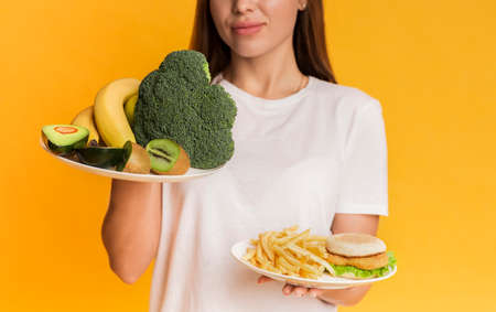 Healthy Vs Unhealthy. Unrecognizable girl choosing between plates with organic fruits and vegetables and junk food, yellow background, croppedの写真素材