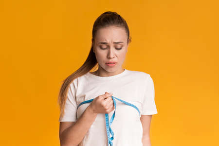 Concerned girl measures her breast with measuring tape on yellow background, checking parameters of chest after dieting, free spaceの写真素材