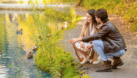 Joyful young lovers feeding birds at autumn park, sitting next to river, panorama with copy spaceの写真素材
